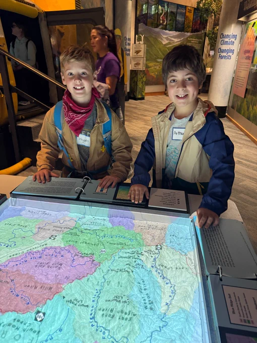 2 fourth grade boys stand in the interactive museum and look at a display that is lighting their faces up underneath their chins