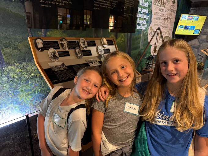 three girls lean their heads together and smile. behind them is the museum and different displays of influential people