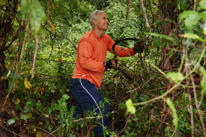 Virginia lacrosse head coach Lars Tiffany in an orange shirt using a manual saw to cut through thick branches in a lush, green forest.