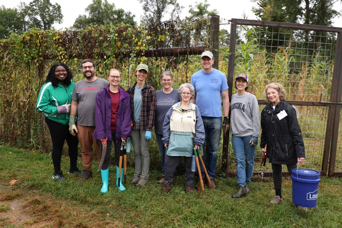 Group of people volunteering at a community garden, standing in front of a metal fence with tools such as shovels and buckets.
