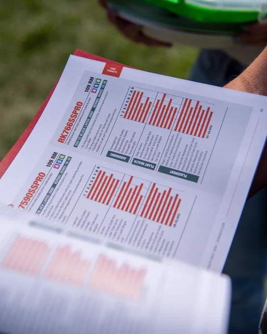 Person holding a 2026 Renk Seed product catalog with pages displaying orange bar graphs and text for each product.