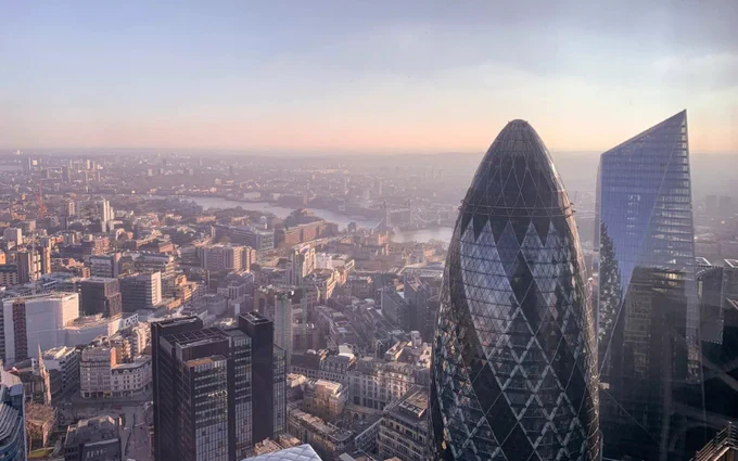 Aerial view of London at sunset featuring the Thames River, the Gherkin, and the Scalpel under a hazy sky.