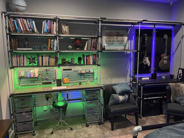 Industrial-style home office setup featuring a Wall Control Metal Pegboard with green backlighting, organized storage bins, bookshelves above, and a workstation below; guitars are displayed on the right side with blue accent lighting.