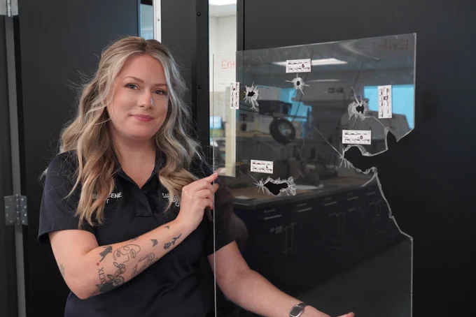Crime scene technician pointing at a piece of glass with multiple bullet holes marked for evidence.