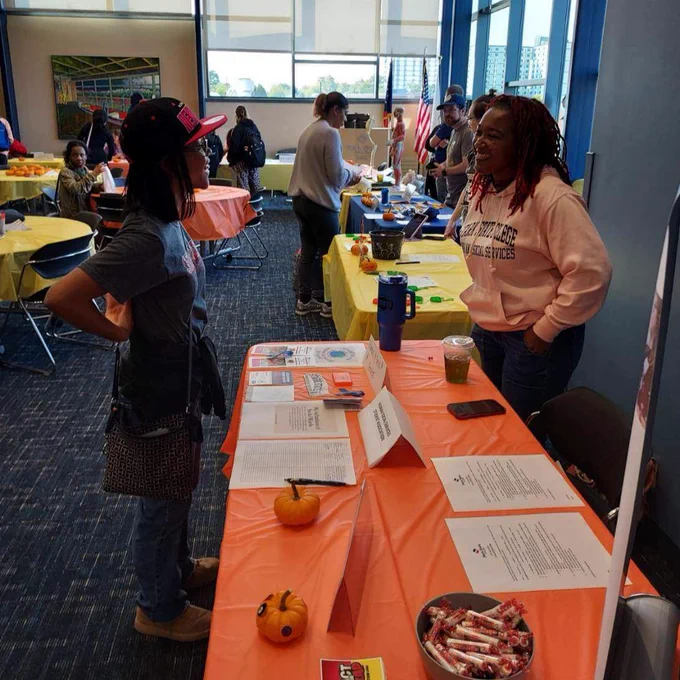 Two individuals engaging in conversation at a Stark State College event at a table decorated with an orange tablecloth, informational pamphlets and small pumpkins. The setting includes other attendees and booths in a room with large windows.