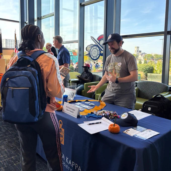 Two people interacting at a Phi Theta Kappa information table, decorated with flyers, candy, and small pumpkins, at a lively indoor event at Stark State College.