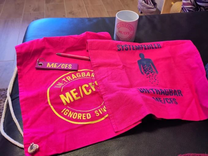Auf einer schwarzen Couch liegen: eine weiße Tasse mit rosafarbener Aufschrift "Ungenießbar ME/CFS ignored since 1969" außerdem einen pinken Turnbeutel mit goldener Aufschrift "Untragbar me-Cfs ignored since 1969". Auf dem Turnbeutel liegt ein rosafarbener Kuli und ein Schlüsselanhänger mit einem lila Stoffstreifen auf dem mit Gold "Me-CFS" steht. Daneben liegt ein pinker Stoffbeutel auf dem in dunkelblau steht "Systemfehler untragbar Me/CFS" außerdem ist eine dunkelblaue Personen abgebildet, die nach unten sich in Pixel auflöst.