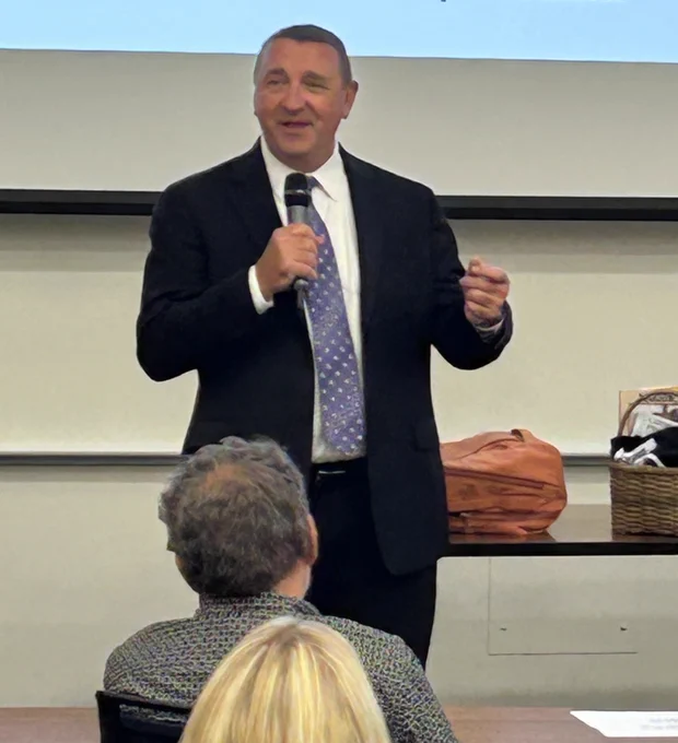 The mayor, a tall white man in a suit with short hair, smiles while holding a microphone.