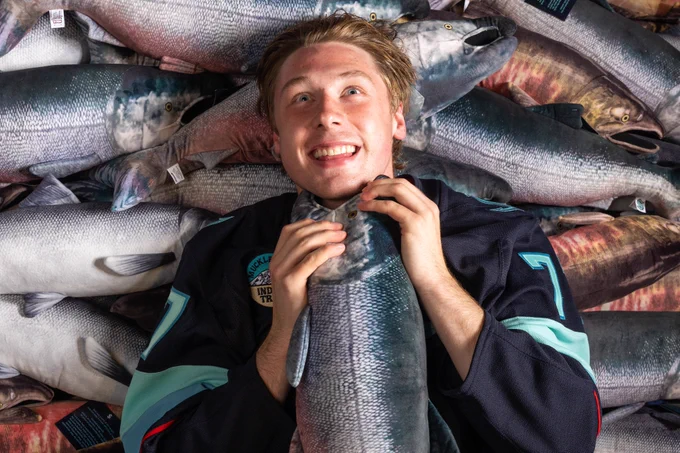 Seattle Kraken prospect Berkly Catton reacts as a king salmon plushie chokes him.