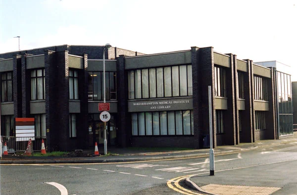 A modern black brick building with large windows, labelled "Wolverhampton Medical Institute and Library," sits on a street corner. A sign indicates a 15-mph speed limit and traffic cones are placed near the entrance. The road in front has yellow markings and a sidewalk with tactile paving is visible.