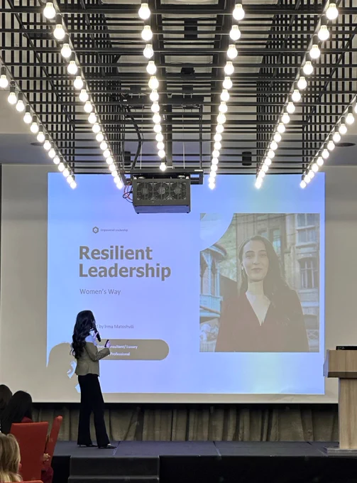 A woman in business attire stands on stage in front of a presentation slide reading 'Resilient Leadership.'