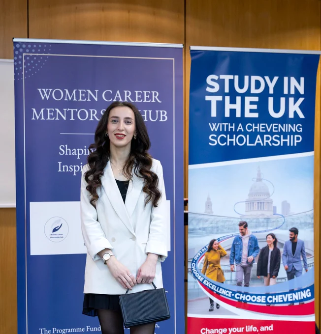A woman in a white blazer stands in front of banners promoting Women Career Mentorship Hub and Chevening scholarships for studying in the UK.
