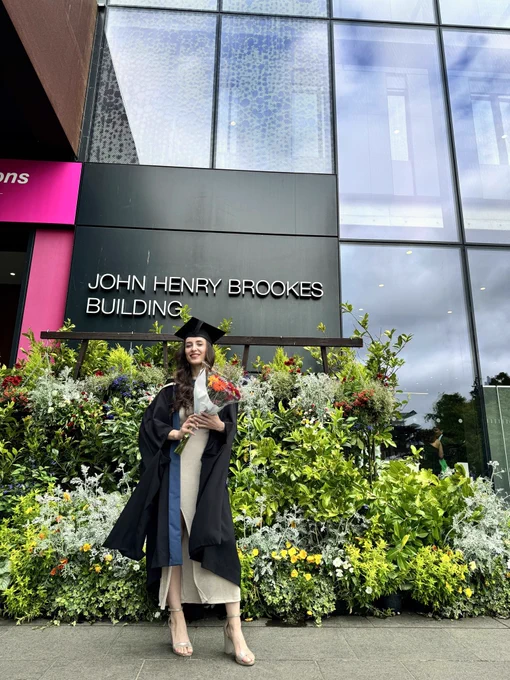 Graduate in cap and gown holding a bouquet, standing in front of the John Henry Brookes Building with lush greenery behind.