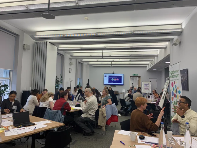 Photo of a large group of  people in a room sat at tables chatting animatedly. 