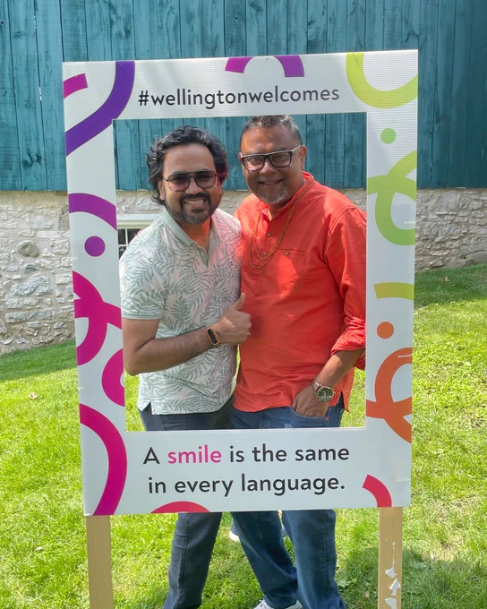 Two men stand together outdoors behind a colorful frame with the hashtag #wellingtonwelcomes and the phrase 'A smile is the same in every language.'