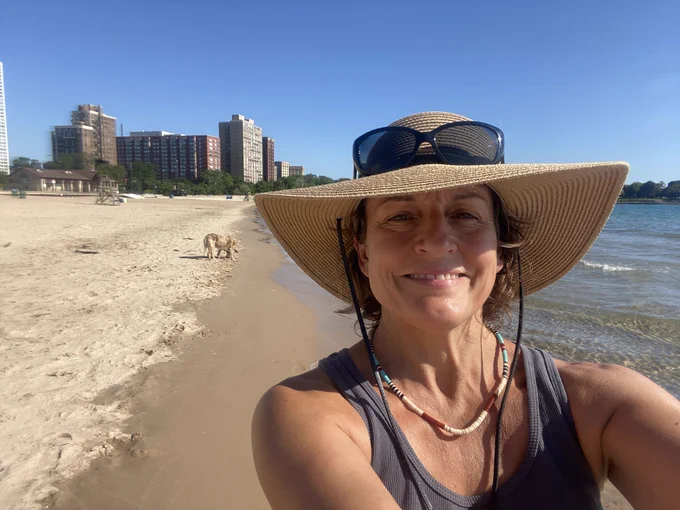 Selfie of a lady wearing a straw hat on the convergence between a sandy beach and waves in front of a city skyline.