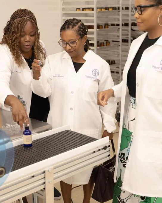 A group of Black women in lab coats inspect a bottle rolling off a production line