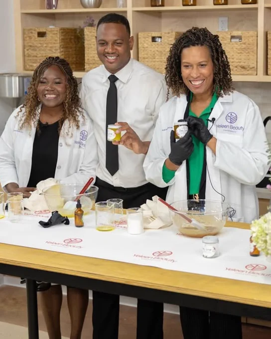 Two black women and a black man show off a table full of cosmetic products, standing behind a counter of lab tools