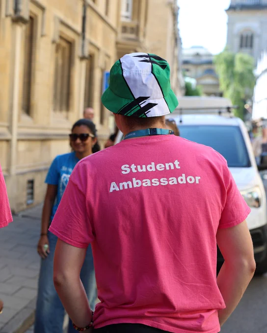 student ambassador wearing bright pink t-shirts with "Student Ambassador" and wearing a green-and-white bucket hat. They’re chatting with another student in a blue t-shirt.