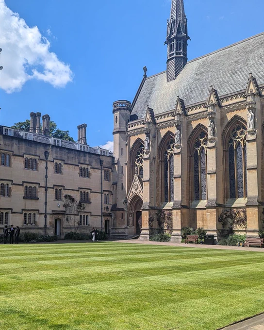 Exeter College’s Front Quad on a sunny summer day. The grass is neatly mown, with the chapel on the right showcasing large Gothic windows and stone statues. A few people are walking in the quad.
