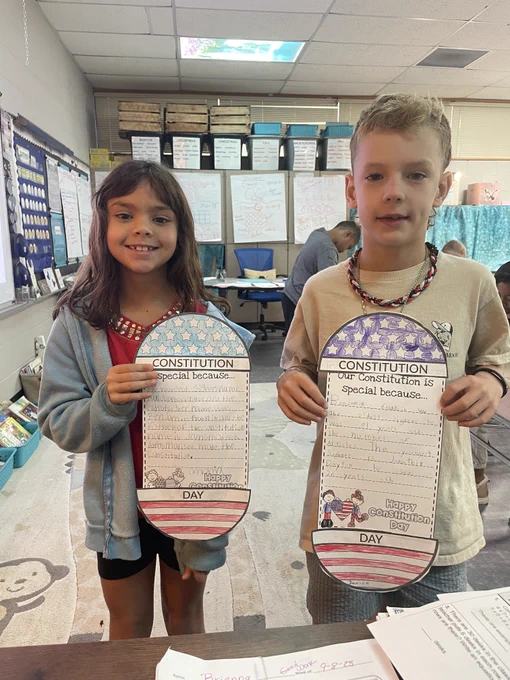 two third grade students (girl in blue jacket, boy in beige shirt) stand holding their papers that read "the constitution" with some writing that they've done 