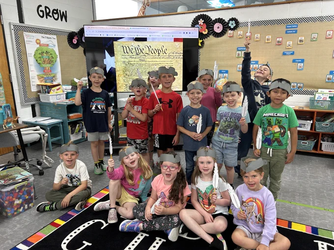 mrs. anders' first graders stand in front of the tv screen with a projected constitution. they are all wearing hats and holding pens