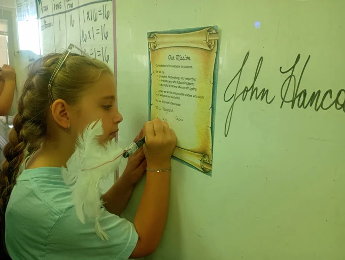 girl with blonde braids and glasses on her head signs a mission statement made to look like the constitution with a white feather pen