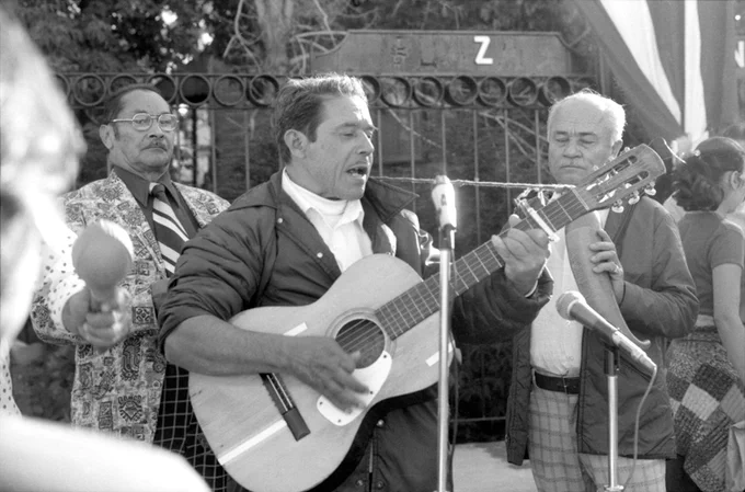 Black and white image of a man playing guitar and singing into a microphone with other men playing instruments behind him