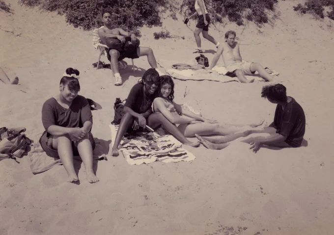 Sepia-toned image of three women sitting on towels at a beach, smiling at the camera. Other people are lounging around them