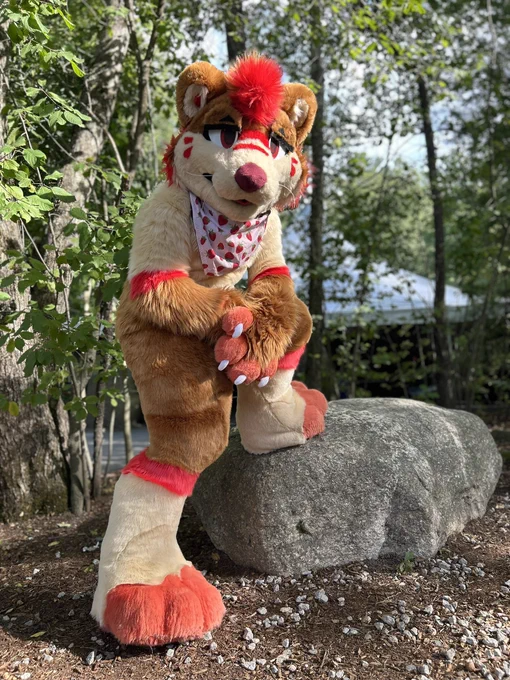 A strawberry themed otter fursuit, one leg up on a rock, the other on the ground. They are seen wearing a strawberry bandana
