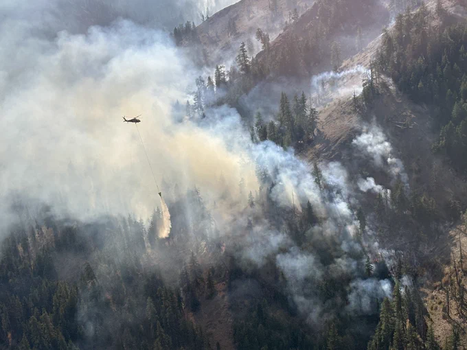 Aerial view of large, smoking wildfire as a helicopter makes a water drop on the burning forest.