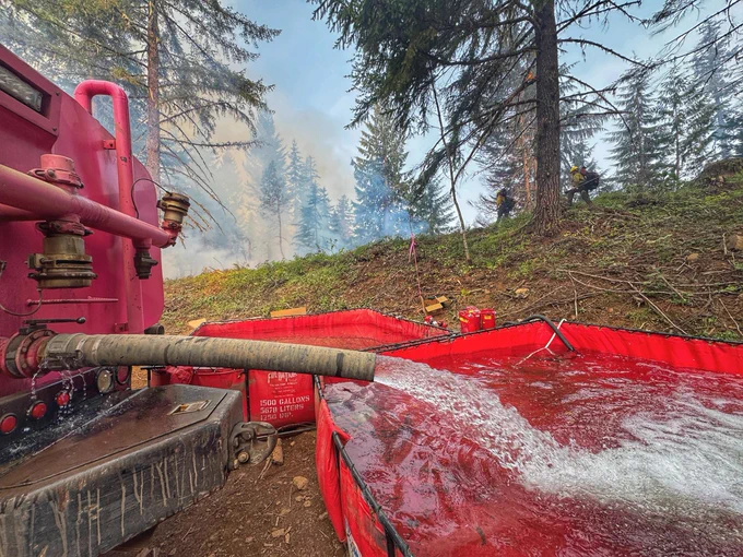 Water flows from a red truck into a portable storage tank, with smoke and firefighters in the background forest.
