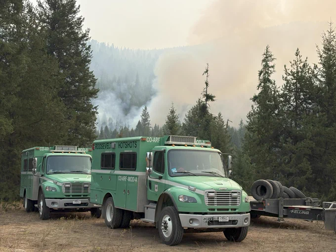 Parked green firetrucks in a forest are close to a burning fire and smoke in the distance. 