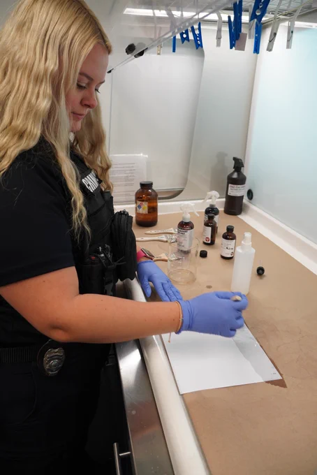 A woman in a police uniform and blue gloves works in a lab setting, applying chemicals to a sheet of paper on a counter with bottles and lab equipment nearby.