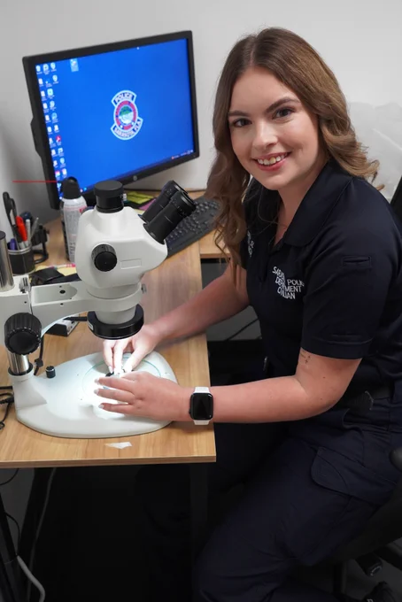 A woman in a police civilian uniform sits at a desk using a microscope. She is smiling at the camera with a computer in the background displaying a police department logo.