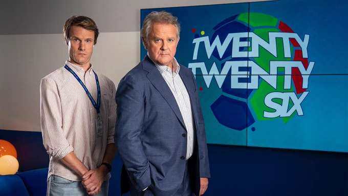 A man in a shirt and jeans and a man in a suit stand by a large screen featuring a football logo and the words Twenty Twenty Six