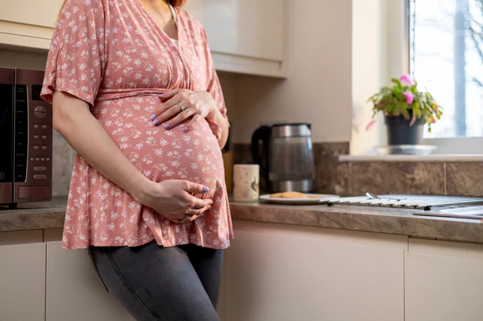 A pregnant woman standing against a kitchen worktop with her hands over her bump.