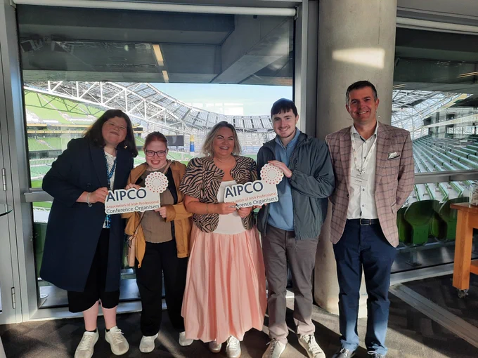 Six people standing together for a photo holding signs saying AIPCO with a view of an empty Aviva Stadium behind them