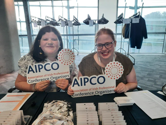 Two TCPID graduates sitting at a conference registration table holding signs saying AIPCO. There are name badges and lanyards on the table in front of them. 