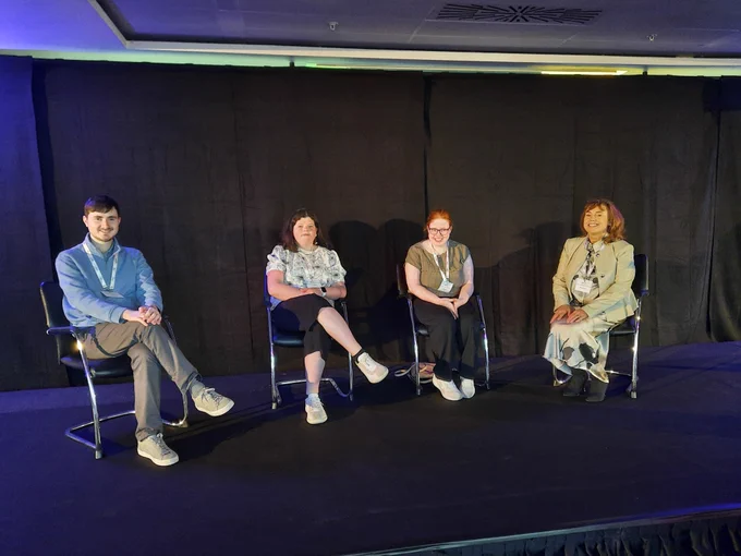 Four people sitting on chairs on a stage at a conference 