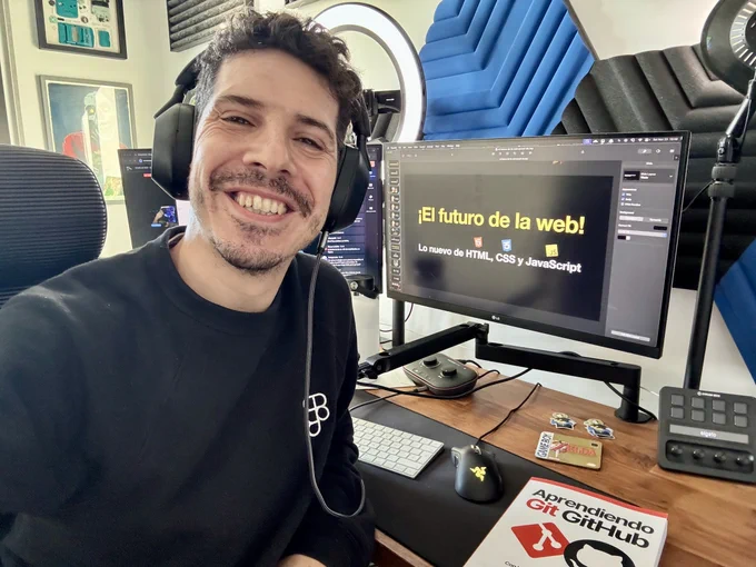 midudev sonriendo a cámara en su setup de programación con una presentación de HTML, CSS y JavaScript en el monitor.