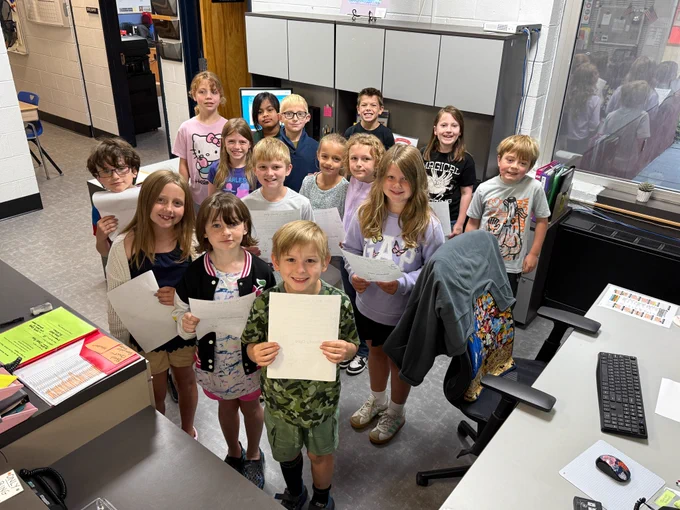 15 third graders stand in the front office in a cluster and smile towards the camera. they are all clutching their papers 