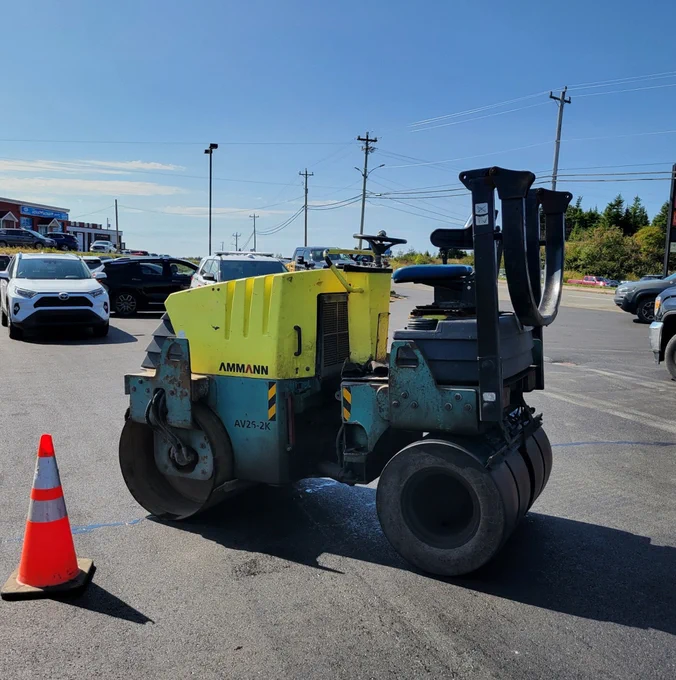 A yellow and green Ammann steamroller, viewed from the side, is stopped next to an orange traffic pylon in a parking lot. No one is on the steamroller and vehicles are visible in the background.