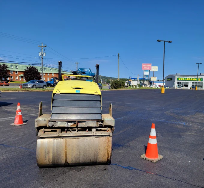 A yellow steamroller, viewed from the front, is stopped between orange traffic pylons in a parking lot. No one is on the steamroller and businesses, and vehicles are visible in the background.