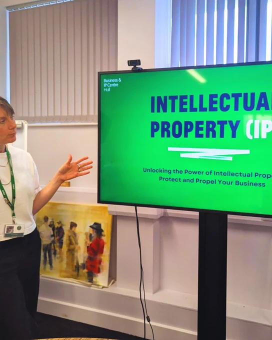 A person presenting a slide titled "INTELLECTUAL PROPERTY (IP)" during a workshop. The slide also reads "Unlocking the Power of Intellectual Property - Protect and Propel Your Business," in a room with paintings on display.