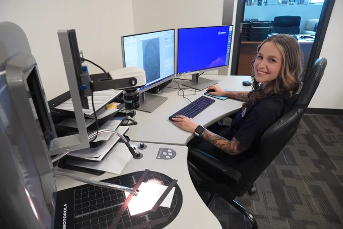 Smiling woman sits at a desk with dual computer monitors, working on fingerprint analysis using specialized forensic software and equipment.