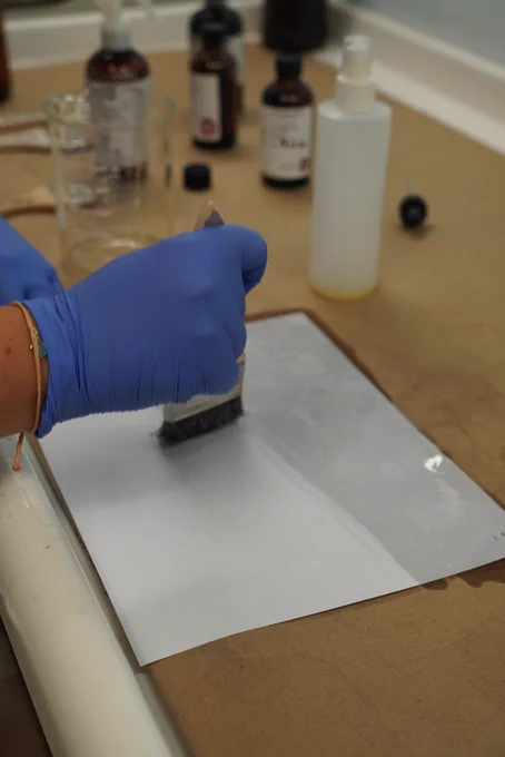 Person wearing blue gloves uses a brush to apply liquid onto a sheet of white paper on a work surface, with laboratory bottles and tools in the background.