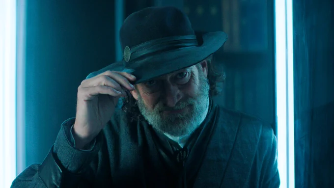 Preem from Foundation is shown with a grey beard and long hair, wearing a dark wide-brimmed hat and futuristic-style coat tips his hat and smiles slightly, standing in a room lit by glowing blue vertical lights.