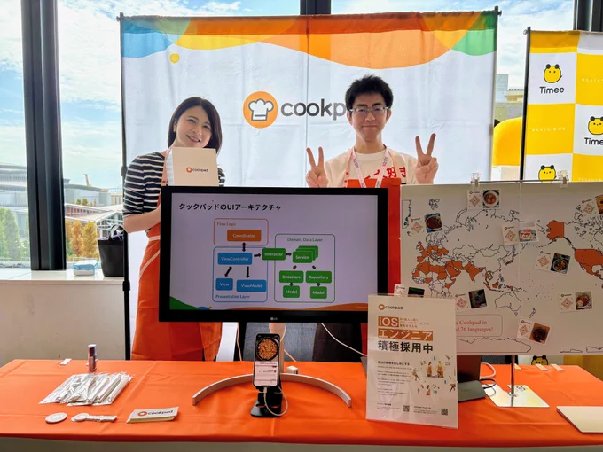 クックパッドのスポンサーブースの様子を写した写真です。ふたりのスタッフが立っており、その前にはアーキテクチャ図やサービス展開国を示す地図などが置かれています。