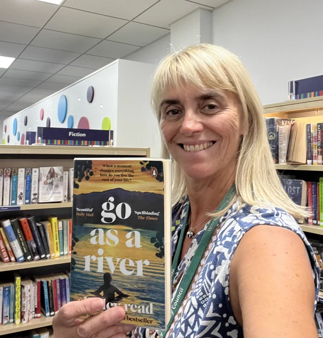Photo of a blond woman who is smiling at the camera and holding up a book called "Go as a River". You can see library book shelves in the background.